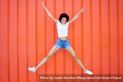 Woman in shorts and t-shirt jumping for joy against outdoor red wall 4dpLD4