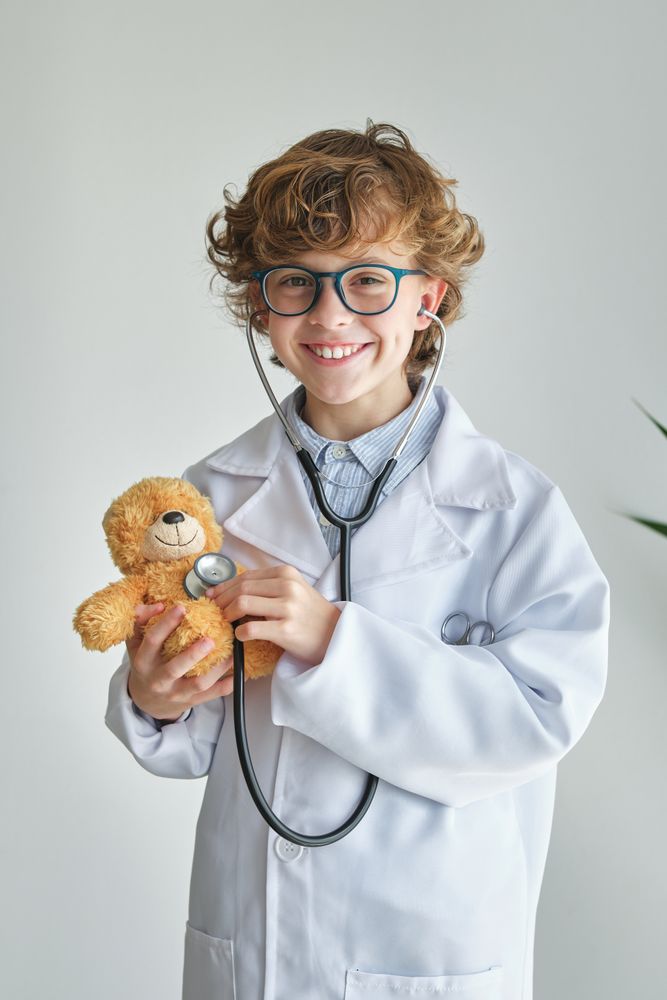 Cheerful boy in medical uniform examining toy bear with stethosc
