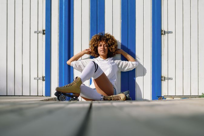 Young Black woman on roller skates sitting in front of a beach hut with hands in her hair