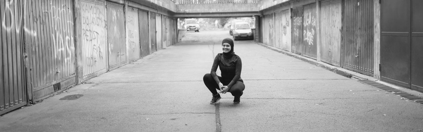 Woman in hijab smiling and crouching outside in a quiet lane
