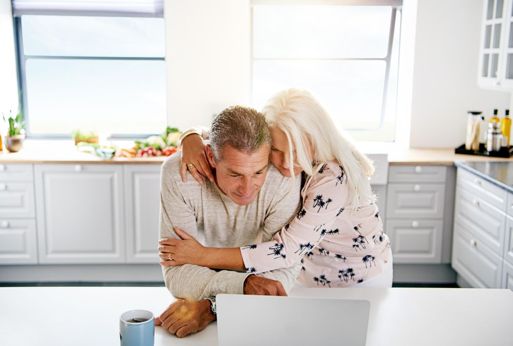 Older couple embracing in front of laptop in bright kitchen