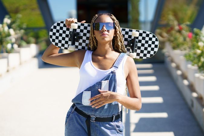 Female in denim overalls with checkered skateboard behind her head with one hand on stomach