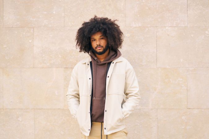 Young man with curly afro hair, dressed in jacket, hands in pockets, leaning against a wall