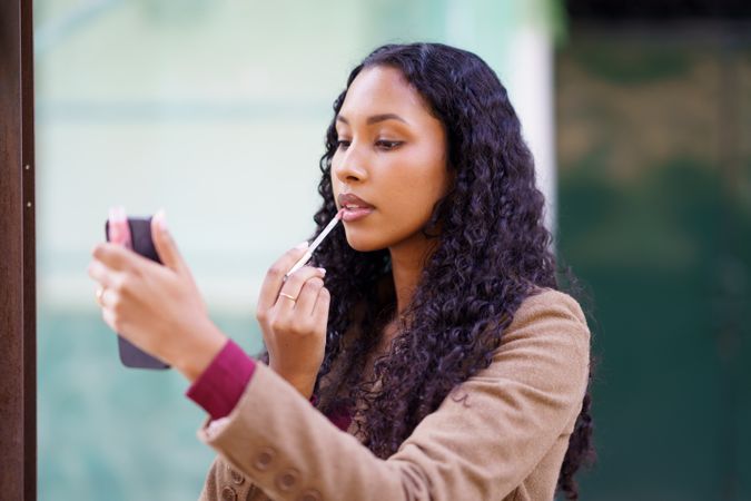 A woman is applying lip gloss while looking at her smartphones mirror, showcasing her beauty