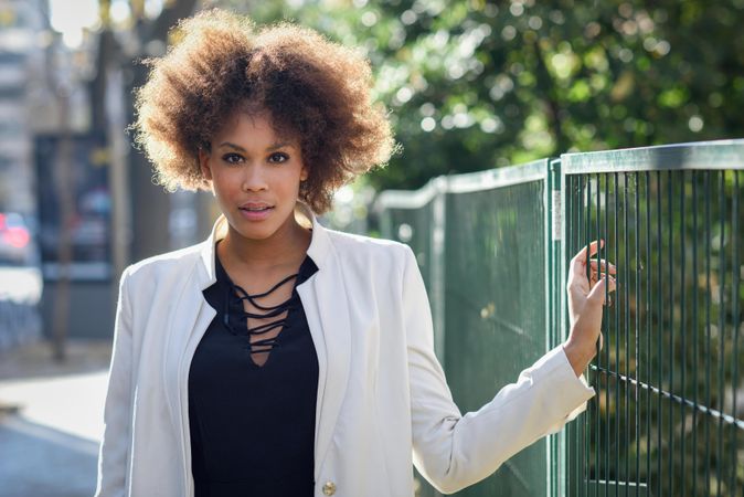 Female with afro looking at camera while standing on street next to green fence