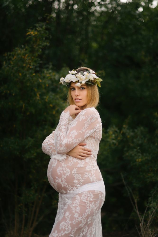 Pregnant woman with closed eyes in white lace dress touching chin while standing in forest