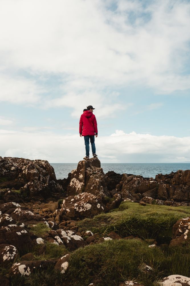 Lonely tourist standing on rocky coast against tranquil sea water under gray sky