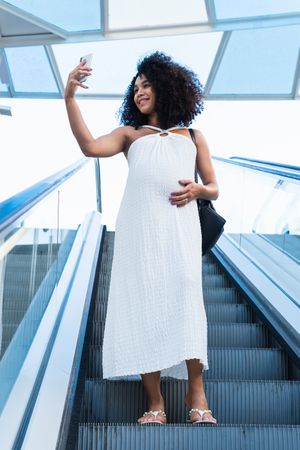 Pregnant Black woman taking self portrait on smartphone on escalator