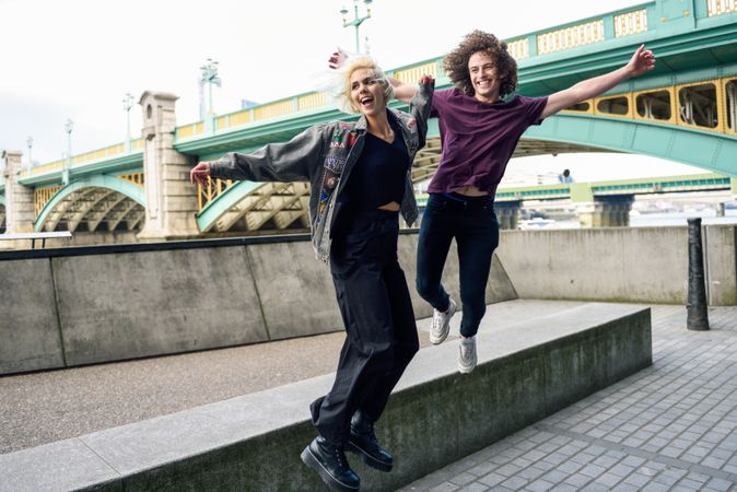 Man and woman jumping and smiling on riverwalk with bridge in background