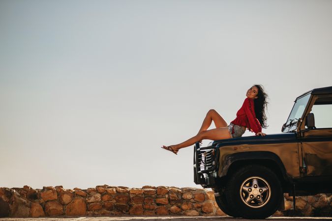 Beautiful young woman sitting on car hood and looking at camera