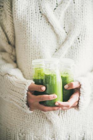 Woman holding three bottles of green smoothie wearing cozy sweater