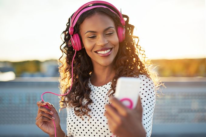 Woman in pink headphones smiling at her phone on rooftop at sunset