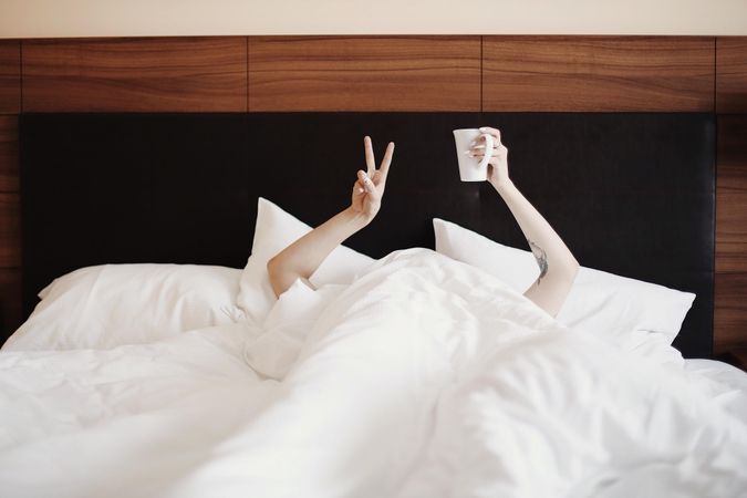 Person under light bed sheets holding ceramic mug and making peace sign