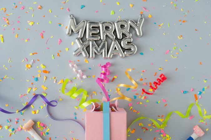 “Merry Xmas” in silver with present with confetti and colorful streamings on grey background