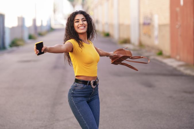Smiling woman spinning around in center of street holding bag and phone