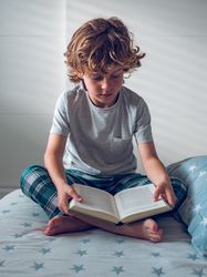 Concentrated little schoolboy reading book on bed