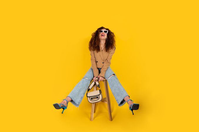 Smiling woman with big hair and sunglasses sitting on chair against yellow background