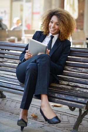 Happy woman in suit laughing at something on her digital tablet on outdoor bench