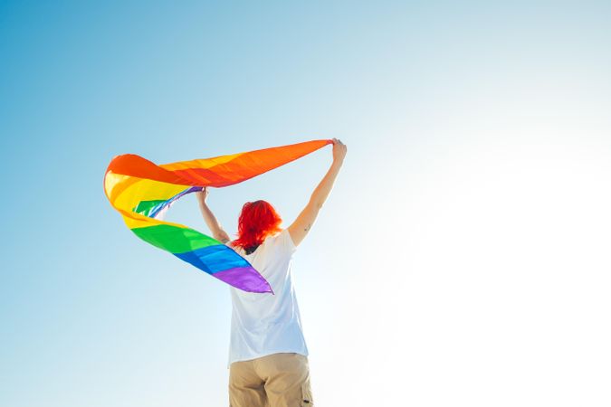 Back view of woman in light top waving rainbow flag under blue sky