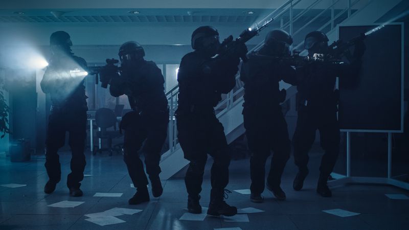 Masked Team of Armed SWAT Police Officers Move in a Hall of a Dark Seized Office Building with Desks and Computers.