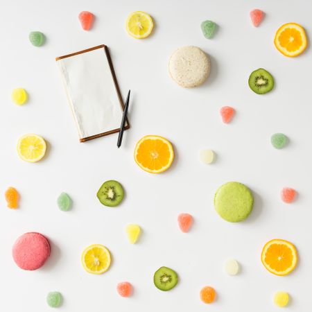 Fruit slices, gummies, and macarons and notepad on light background