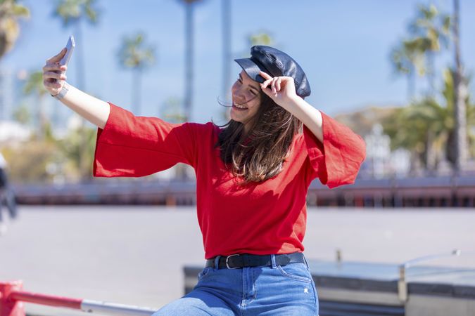 Smiling young woman casual clothing sitting on a metallic fence while taking a selfie on a bright day