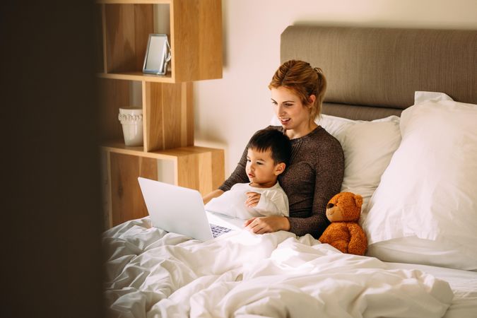 Young mother with her beloved son in the bedroom on the bed watching a movie on laptop