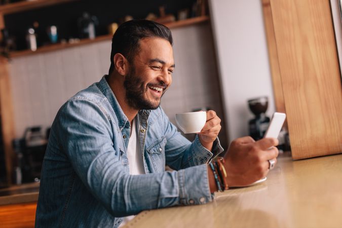 Smiling young man sitting at cafe using mobile phone and having coffee