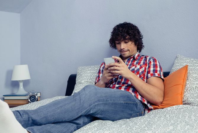 Young man with curly hair looking smartphone in a bed