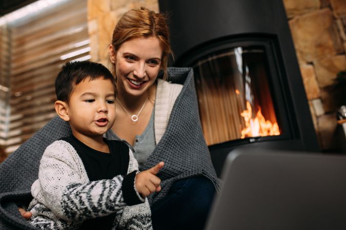 Little boy pointing at laptop while sitting with mother near fireplace at home