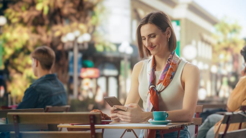 Portrait of a Happy Female Using Smartphone on a Restaurant Terrace, Enjoying a Cup of Coffee. Young Woman Smiling When Receiving a Text, Chatting with Friends Online, Checking Social Network Updates