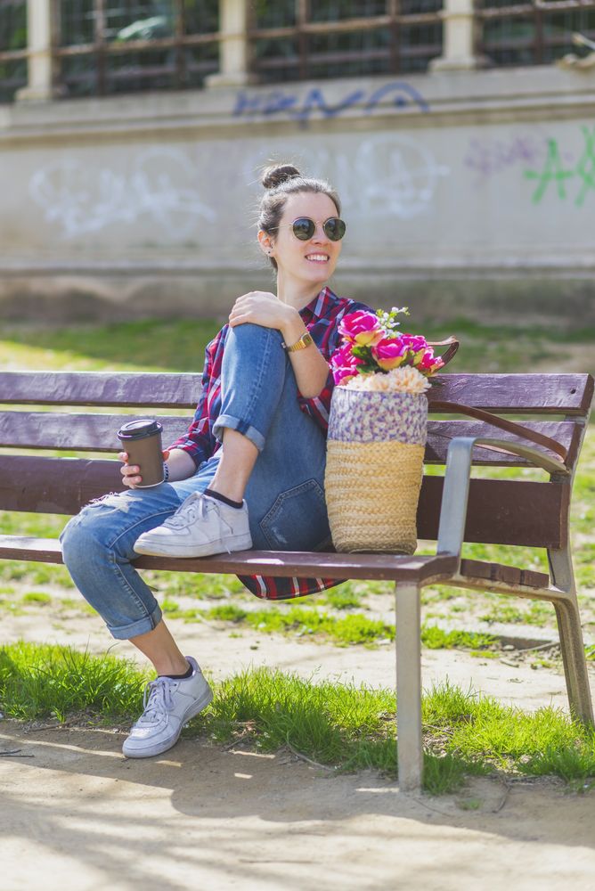 Front view of a young hipster woman sitting on a park bench relaxing in a sunny day while looking away
