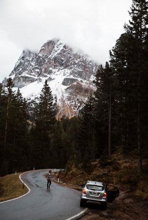 Lonely road with car on roadside and traveler among pine forest near big cliffs