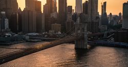 Iconic New York City Landscape Over East River with Skyscrapers, Brooklyn Bridge, Cars and Ferry Boats. Cinematic Evening Urban Skyline with Sunset and Lightly Clouded Sky