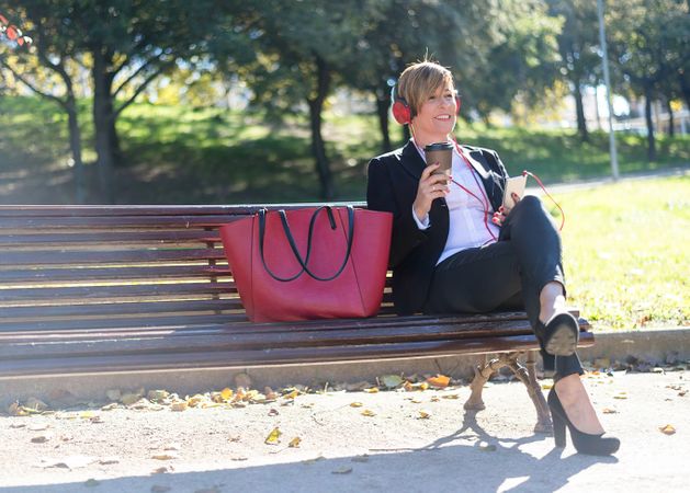 Professional woman sitting on park bench enjoying coffee and listening to music