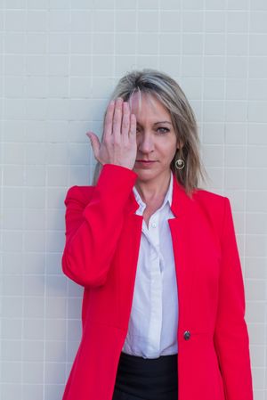 Portrait of a blonde woman wearing red jacket against a tiled wall covering her eye