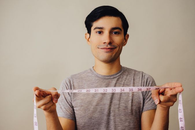 Hispanic male holding measuring tape between hands in beige studio shoot and looking at camera