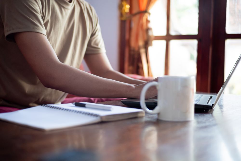 Asian Male with shirt working from home in living room with book, laptop and cup of coffee.