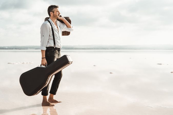 Focused stylish man with suitcase and guitar gig bag on seaside