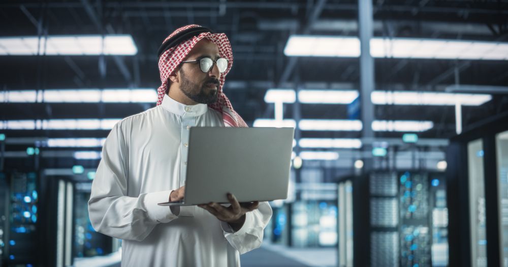 Portrait of a Middle Eastern IT System Engineer Wearing Traditional Robe, Using Laptop Computer in a Modern Technological Server Farm Facility.