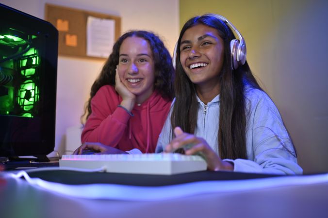 Cheerful teenage diverse girlfriends sitting at table with gaming PC and playing game