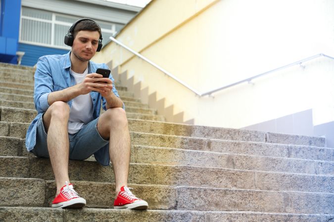 Young man in headphones sits on the steps and looking in phone
