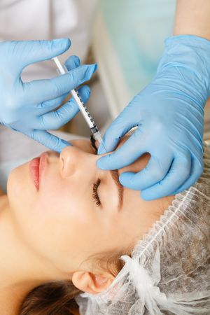 Cosmetologist at med spa injecting botox into forehead wrinkles in female client, vertical