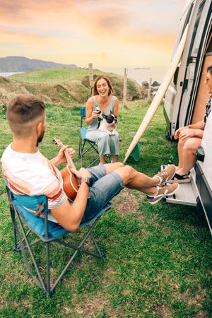 Three friends sitting outside a motorhome with beers, a ukulele and a dog, vertical