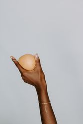 Black hand holding a pear wearing a gold bracelet against pastel blue background