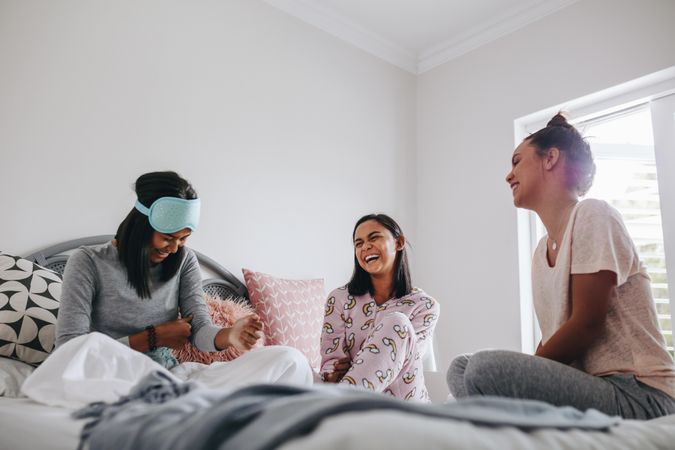 Young women having fun during sleepover sitting on bed and laughing