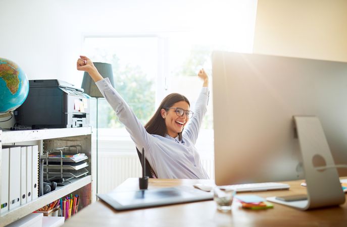 Smiling female sitting at her desk with her hands up in the air in celebration