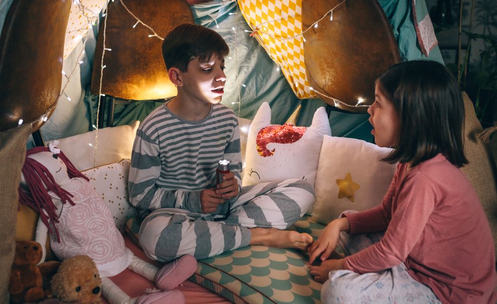 Boy illuminated with lantern telling a terror story to surprised girl sitting in dark living room