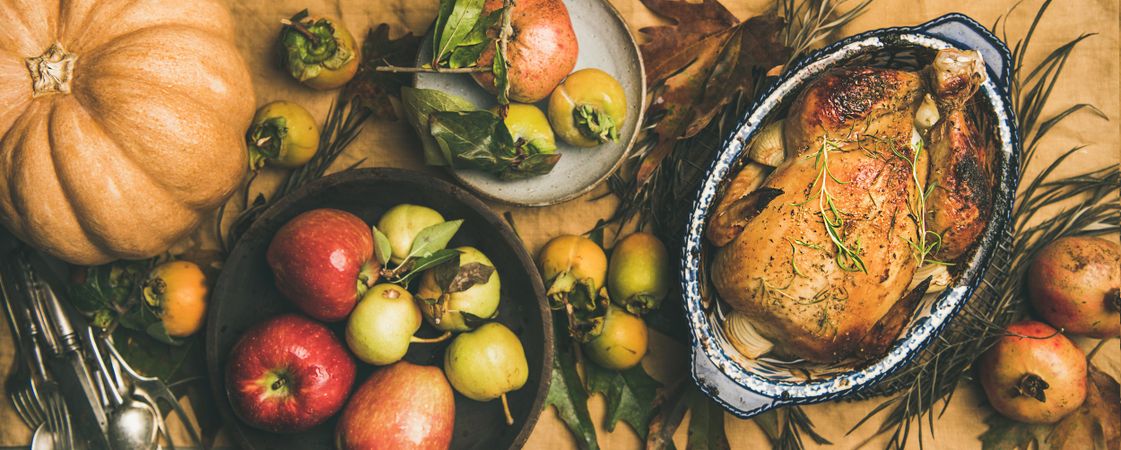 Roast turkey in decorative roasting pan, on table with fall leaves and fruit, wide composition