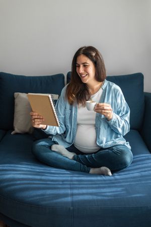 Cheerful pregnant woman drinking beverage on sofa at home